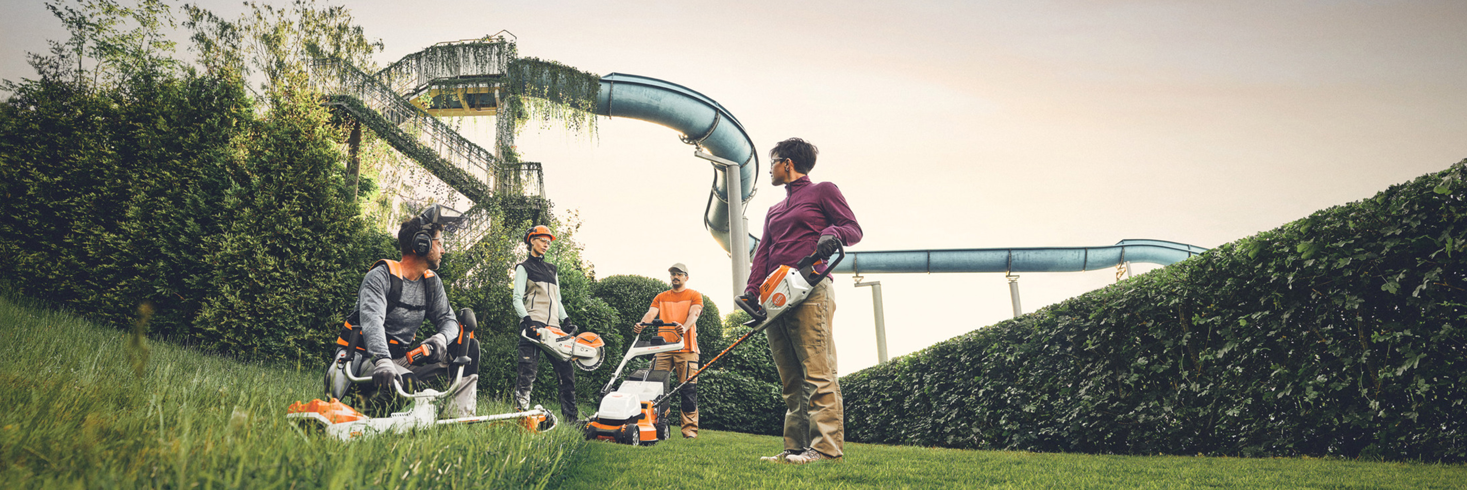 Група луѓе работат со градинарски алатки STIHL во зелена површина со голема жива ограда и водни тобогани во позадина. Тие се опремени со тримери, косилки за трева и заштитна опрема.