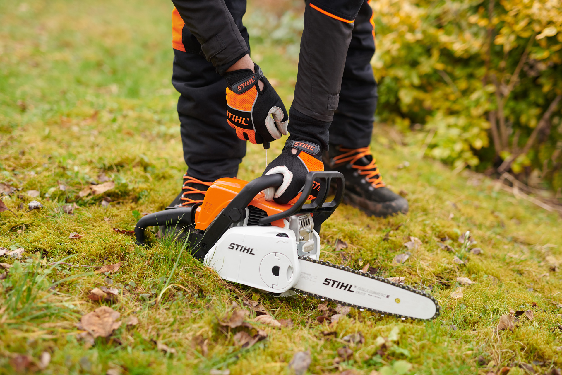 Моторна пила STIHL MS 180 C-BE на земја, со лице кое носи заштитни ракавици како ја држи рачката.