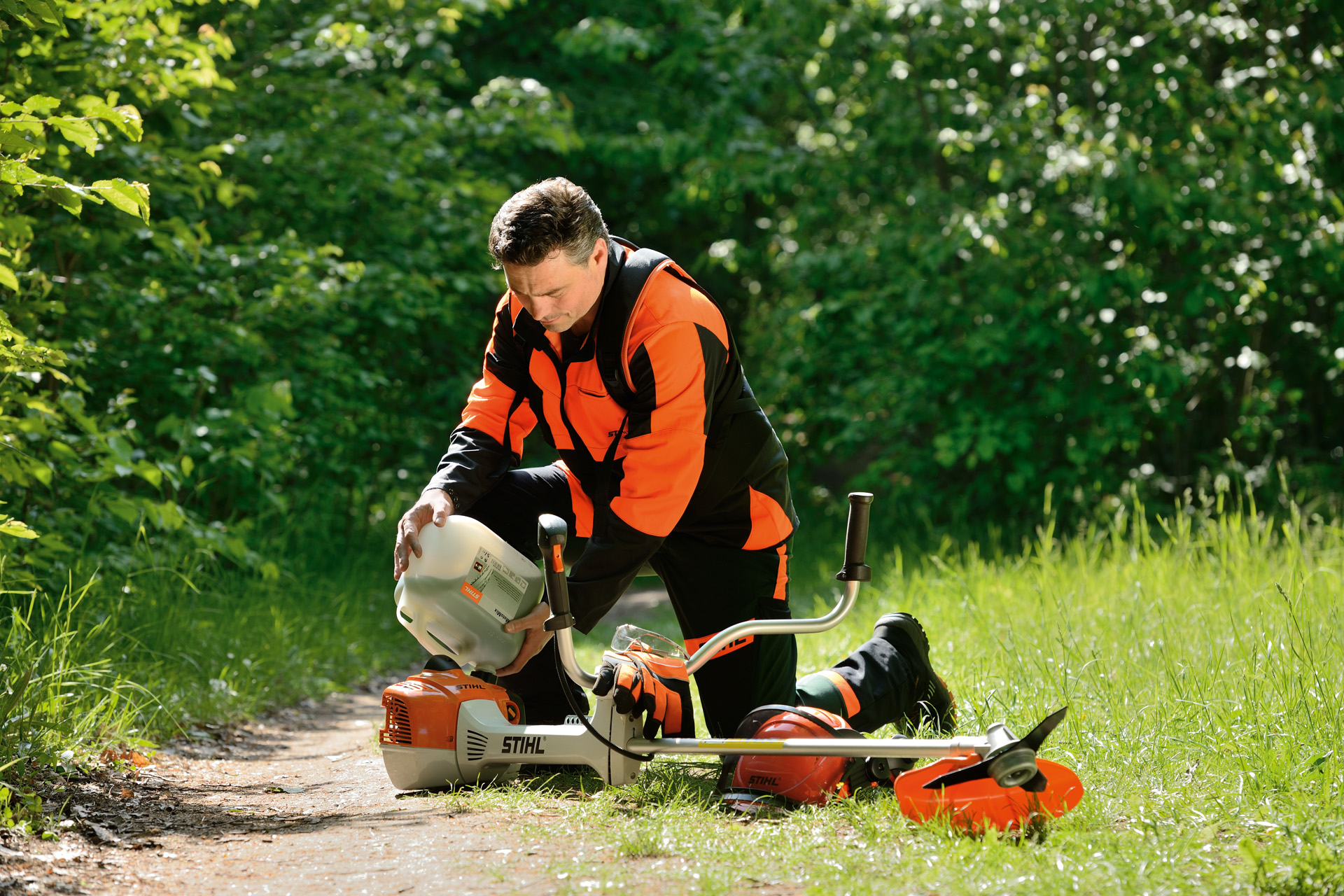 Човек го полни резервоарот за гориво на тримерот за трева STIHL од големо шише со грмушки зад него