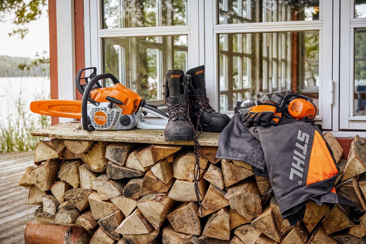 Заштитна облека STIHL и MS 162 што лежи на полица над наредени дрва за огрев