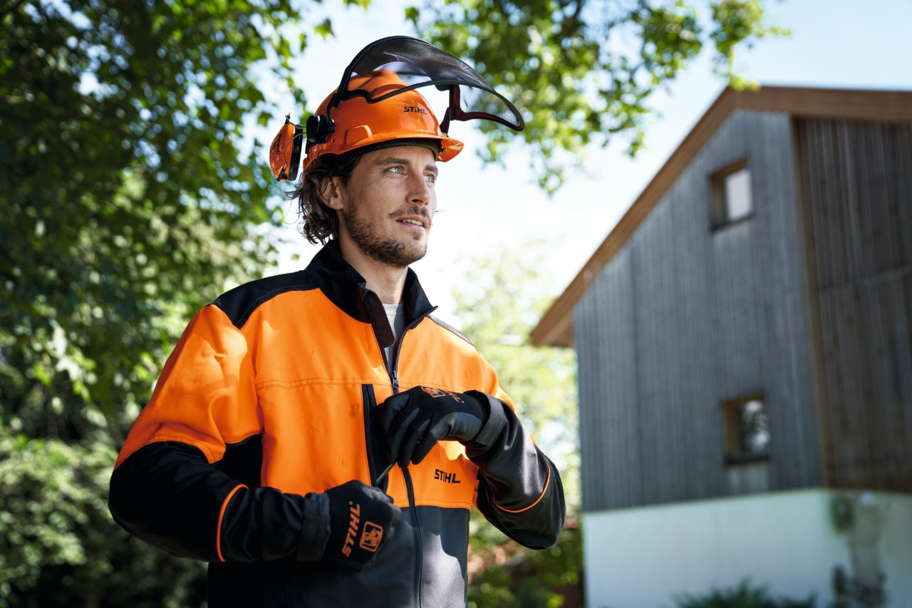 Човек облечен во портокалова и црна заштитна опрема STIHL, вклучувајќи кацига и ракавици, самоуверено гледа надвор од сликата, со куќа и дрвја во позадина 