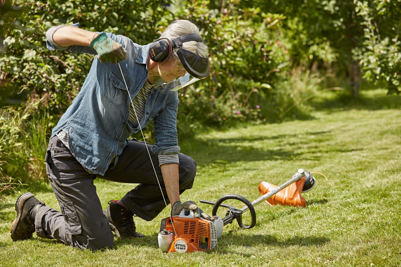 Жена носи заштита за уши и лице и стартува тример STIHL FS 56 R на тревна површина.