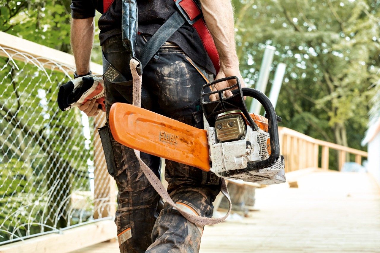 Бензинска моторна пила STIHL MS 261 со штитник за ланецот ја носи маж додека преминува мост.